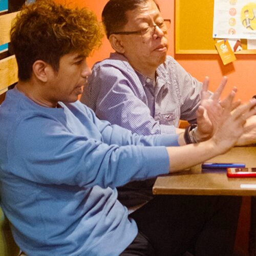 About Us 1 Francis and his teammate are seated at a table in a casual setting, engaged in conversation